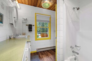 Un baño con ventana de marco amarillo, un lavabo, un espejo, una ducha y suelo de madera.