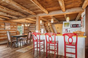 Interior rústico de una cabaña con una mesa de comedor de madera, sillas rojas, una cocina con microondas y una escalera.