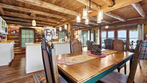 Interior rústico de una cabaña con una mesa y sillas de madera, una zona de cocina y grandes ventanales con vistas a un cuerpo de agua.