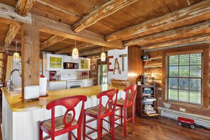 A rustic kitchen with wooden beams, a central island with bar stools, a microwave, and a window with blinds.