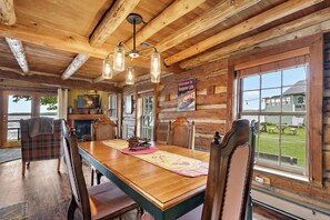 A wooden cabin interior with a dining table, chairs, a fireplace, and large windows.