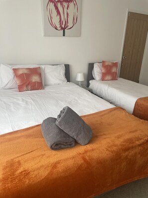 A hotel room with two beds, white bedding, and red patterned pillows. There is a grey towel on the bed and a wooden door in the background. - Immaculate 3-bed House in Luton (Luton)