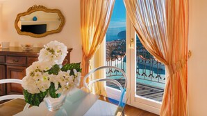 A room with a dining table, white flowers, a mirror, and a view of a coastal area through a window with curtains. - Villa Grazielluccia with Sea View and Pool (Priora)