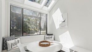 Une pièce lumineuse et ensoleillée, avec une table ronde, des chaises blanches et un banc agrémenté de coussins sur lesquels est écrit « BIENVENUE ».