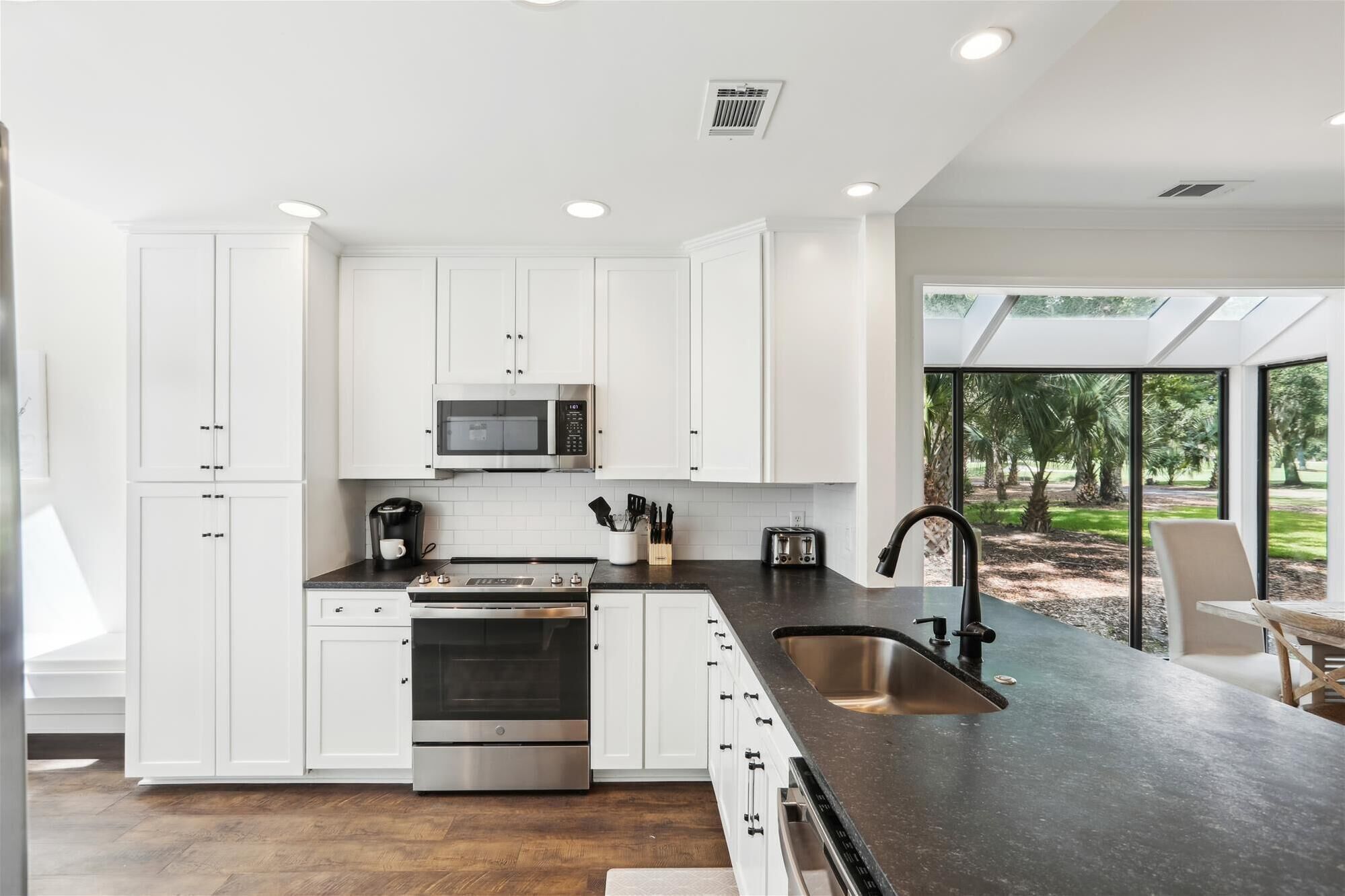 Una cocina moderna con armarios blancos, electrodomésticos de acero inoxidable y un amplio ventanal con vistas a un jardín.