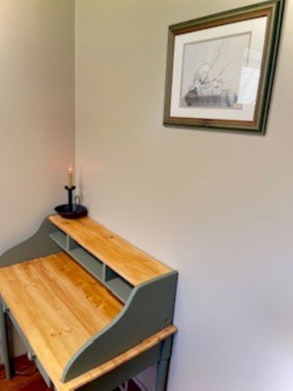 A corner desk with a wooden top and grey legs, a framed picture on the wall, and a lit candle on a stand.