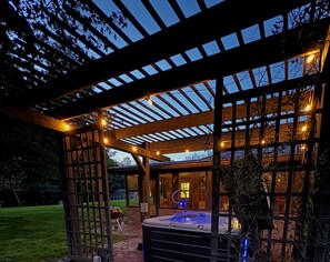 A covered outdoor area with a hot tub, wooden pergola, and a view of a building with large windows. - Secluded house built into the earth. Pet friendly, fishing, hot tub, Wi-Fi! (Magnolia)