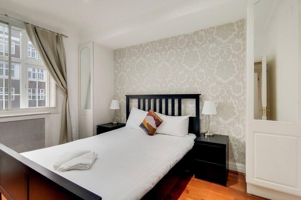 Un lit bien fait, recouvert d’une couette blanche, deux tables de chevet avec des lampes, un papier peint à motifs et une fenêtre avec des rideaux.