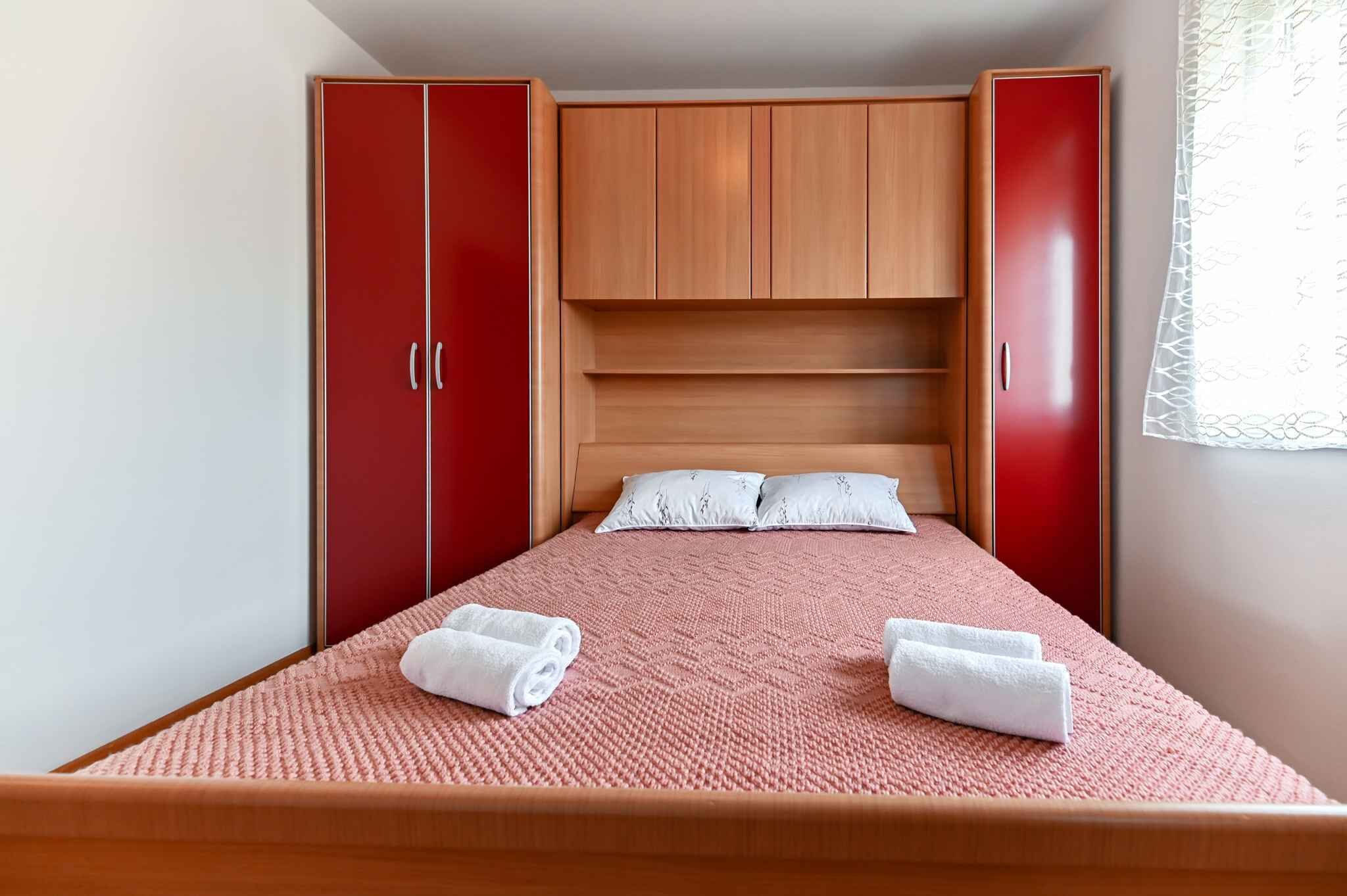 A bedroom with a bed, red and wooden wardrobe, and a window with a white curtain.