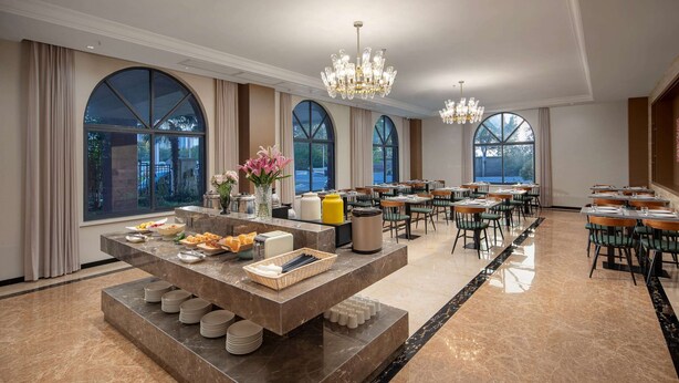 Un amplio comedor con una mesa buffet central, varias mesas de comedor y grandes ventanales con cortinas.