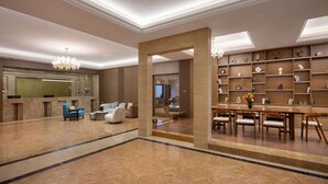 Un vestíbulo amplio con suelo de mármol, una recepción, una zona de estar con sofás y sillones, un comedor con mesa y sillas de madera, y una pared con estantes que exhiben diversos objetos.
