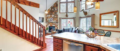 Une cuisine spacieuse avec un grand îlot central, un escalier en bois, une cheminée en pierre et de grandes fenêtres.