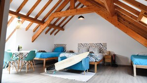 A loft-style bedroom with exposed wooden beams, a dining area with teal chairs, and a surfboard on the floor. - Go4surf Beachlofts (Peniche)