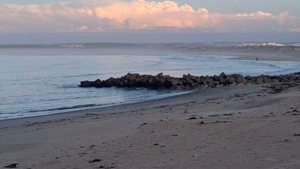 A beach with a rocky breakwater and a distant shoreline with buildings. - Go4surf Beachlofts (Peniche)