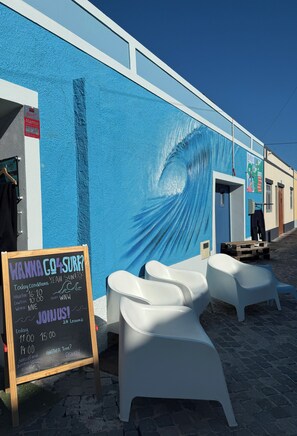 A building with a blue mural, outdoor seating, and a chalkboard sign with surf conditions. - Go4surf Beachlofts (Peniche)