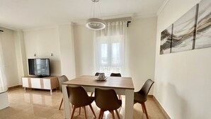 A dining area with a wooden table, four chairs, a television on a cabinet, and a window with curtains. - Alq 292 Porto Azul 14 (Calpe / Calp)