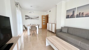 A living room with a TV, a sofa, a coffee table, a dining area with chairs, and a door leading to another room. - Alq 292 Porto Azul 14 (Calpe / Calp)