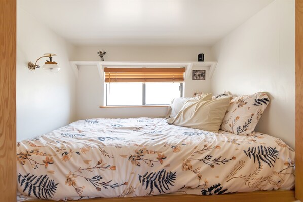 A bedroom with a bed, floral bedding, a window with blinds, a wall-mounted light fixture, and a small shelf with items. - The Desert View Cabin (Monticello)