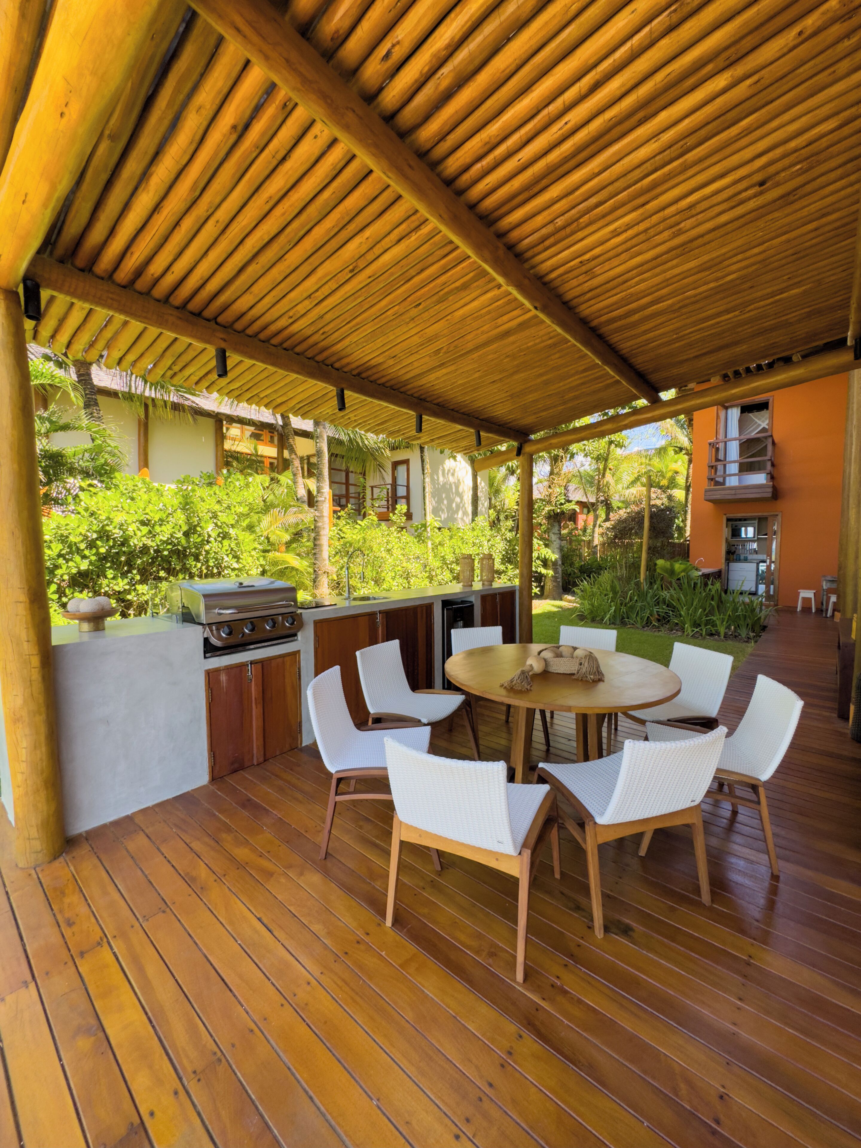An outdoor patio with a wooden deck, a round table with white chairs, a grill, and a small kitchen area.