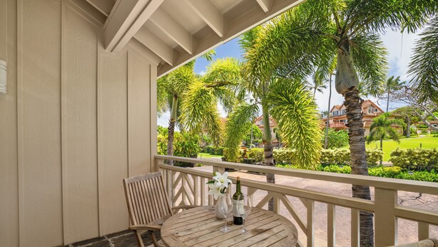 A wooden table with two chairs and a vase with a flower, set on a balcony with a view of palm trees and a building in the background.