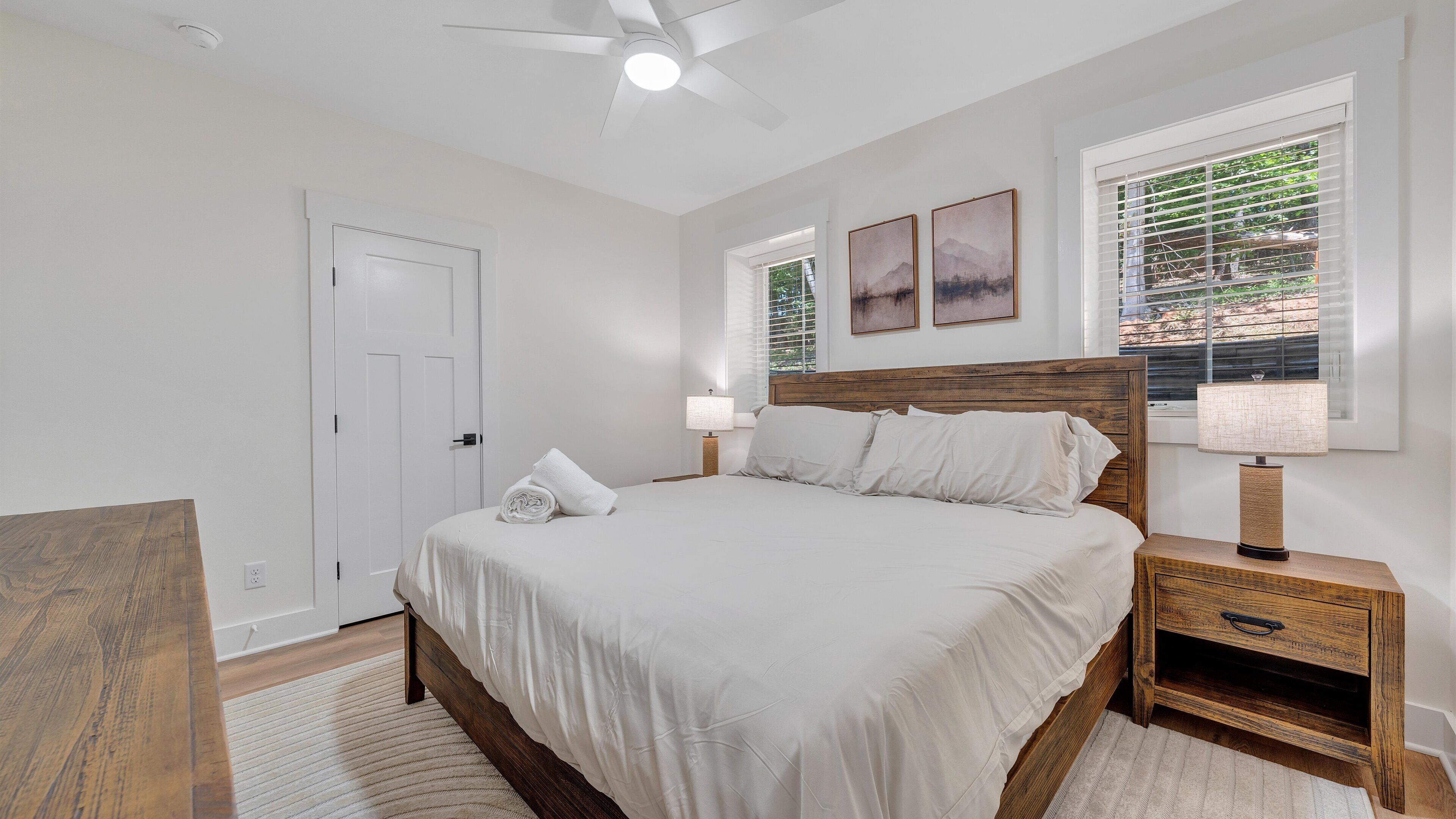 A bedroom with a large bed, a nightstand with a lamp, a wooden headboard, and a window with blinds.