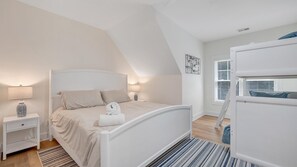 Un dormitorio con cama, mesitas de noche, lámpara, toalla enrollada, ventana con persianas y una cama litera.