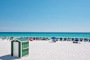 A beach with a green and white beach hut, blue umbrellas, and people relaxing on the sand.