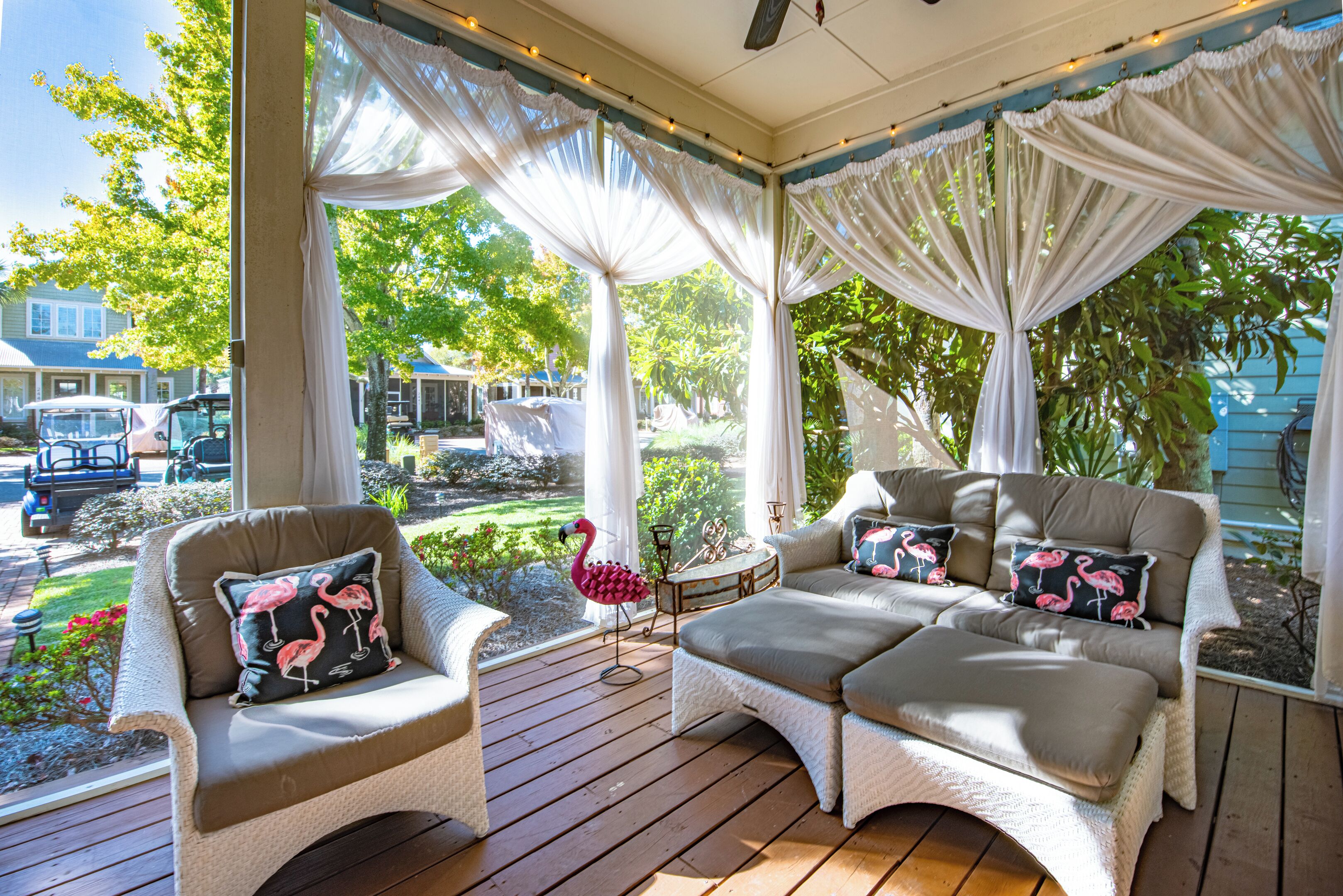 A covered outdoor seating area with cushioned chairs and ottomans, a flamingo sculpture, and a view of a garden and houses in the background.