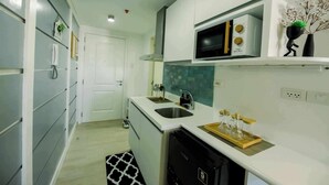 A compact kitchen with a microwave, sink, and a small refrigerator. There is a decorative plant on the counter and a black and white rug on the floor. - Ethan's Crib at Azure North (San Fernando)
