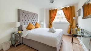 A modern bedroom with a tufted headboard, a glass desk, and a view of the city through orange curtains. - The Portman Residences (London)