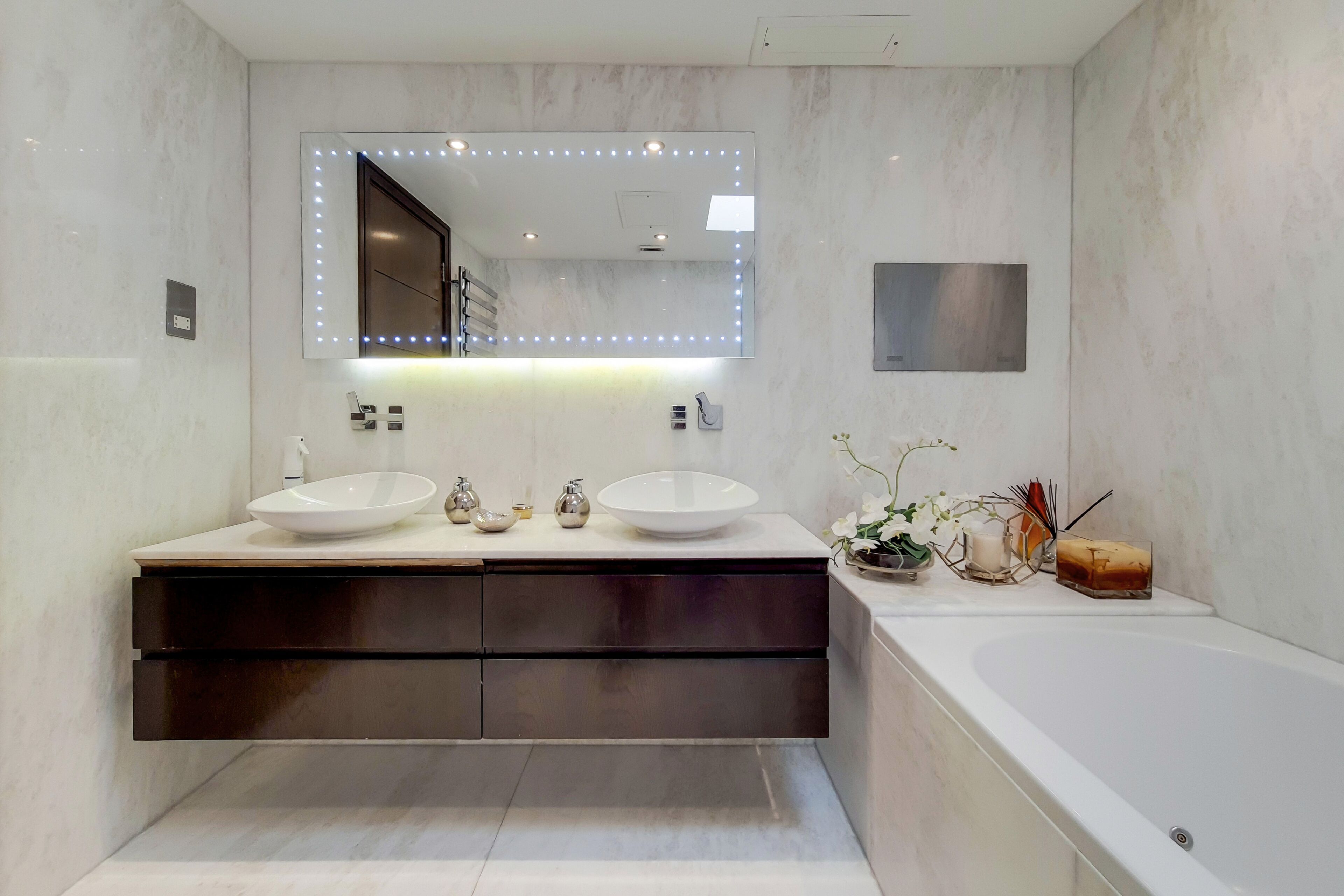 A modern bathroom with two sinks, a large mirror with LED lights, a bathtub, and a dark vanity.