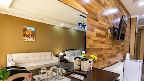 A hotel room with a wooden accent wall, a white sofa, a dining table with a tray of glasses, and a small refrigerator. - Hotel Dimasi (Kutaisi)