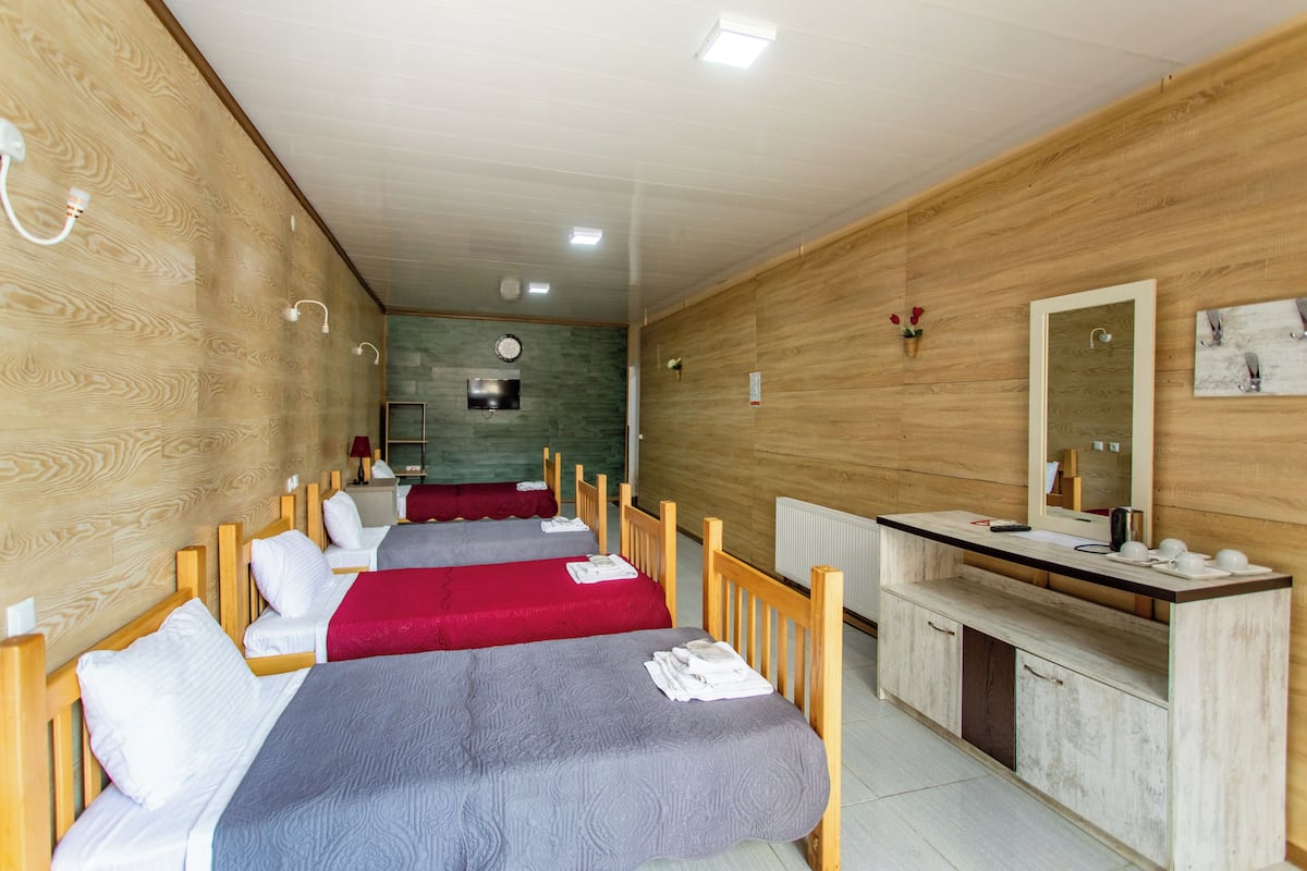 Habitación de hotel con paredes de madera, dos camas con ropa de cama roja y gris, un banco de madera, un espejo y una zona de baño pequeña.