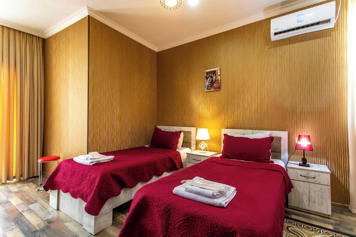 Habitación de hotel con dos camas, cada una con colcha roja, una mesita de noche con lámpara y un equipo de aire acondicionado en la pared.