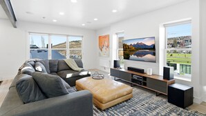 A modern living room with a grey sectional sofa, a tan ottoman, a flat-screen TV, and a large window offering a view of a mountainous landscape.