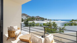 A balcony with a wooden chair and cushions, a view of the sea, and buildings in the background. - Amazing Beach View , Walk to the Beach. Cleaning & Concierge (Cabo San Lucas)