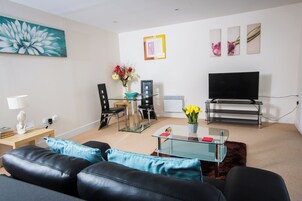 A modern living room with a black leather sofa, a glass coffee table, a TV, and a dining area with chairs and a vase of flowers.