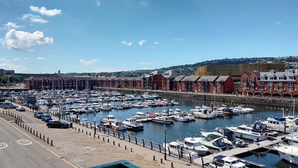 A marina with numerous boats docked, a road with speed limit signs, and a red brick building with a clock tower in the background. - Meridian Tower Apartment (Swansea)
