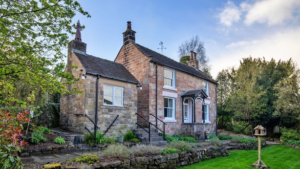 A stone house with a traditional design, featuring a front entrance with steps, a chimney, and a small garden area with a bird feeder. - Spring Cottage (Endon)