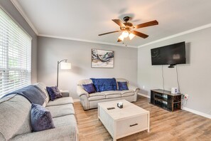 Una sala de estar con un ventilador de techo, un televisor de pantalla plana montado en la pared, un sofá con cojines azules y una mesa de centro con controles remotos.