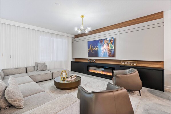 A modern living room with a sectional sofa, a coffee table, leather armchairs, a flat-screen TV, and a fireplace.