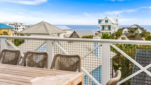 A balcony with wicker chairs and a wooden table overlooking a coastal area with houses and the sea. - Loaded Designer Home with Gulf and Lake Views, Elevator, Heated Pool, and More (Santa Rosa Beach)