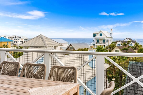 Un balcon agrémenté de fauteuils en osier et d’une table en bois, offrant une vue sur une zone côtière avec des maisons et la mer.