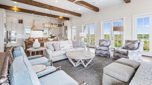 A spacious living room with a large sofa, a coffee table, and a dining area with wicker chairs. There is a kitchen in the background with white cabinets and a large window. - Loaded Designer Home with Gulf and Lake Views, Elevator, Heated Pool, and More (Santa Rosa Beach)