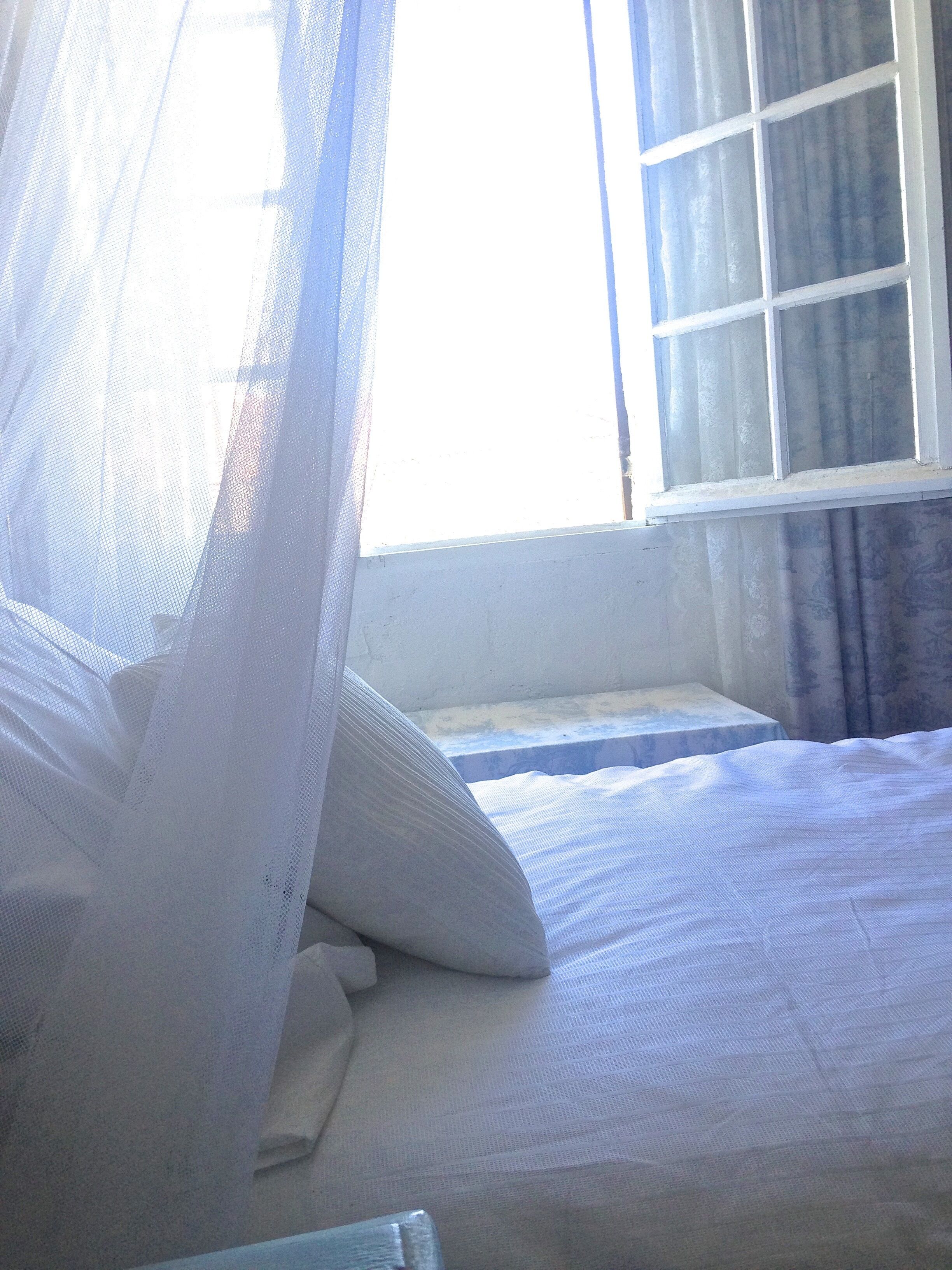 Una cama con ropa blanca, una cortina translúcida y una ventana que da a vistas del exterior.