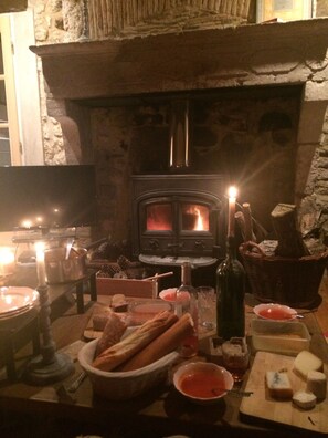 Une pièce chaleureuse avec une cheminée en pierre, une télévision et une table où sont disposés du pain, du fromage et des boissons.