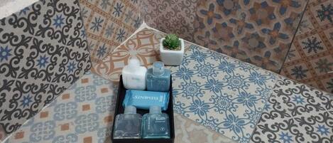 A corner of a room with patterned tiles, a small potted plant, and a tray with toiletries including body wash, shampoo, and bath gel.