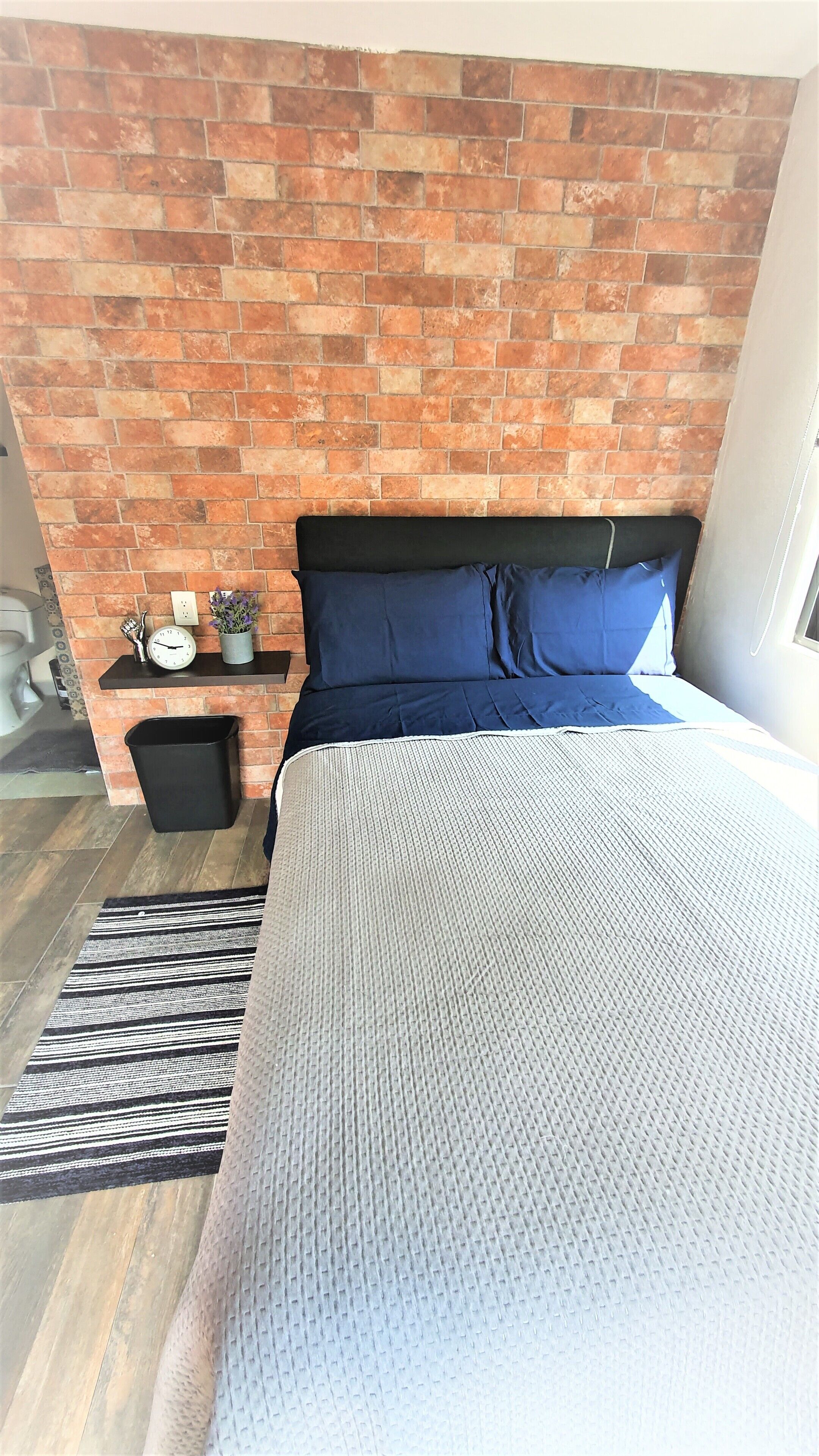 Un dormitorio con pared de ladrillo, una cama con ropa de cama azul, una mesita de noche pequeña con un reloj y una planta, y un cubo de basura negro.