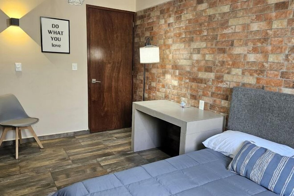 A bedroom with a brick wall, a bed with blue bedding, a desk with a lamp, and a motivational quote on the wall.