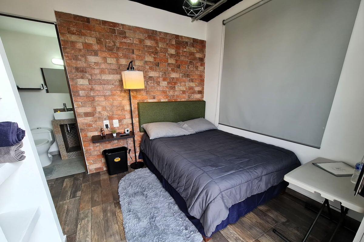 Een slaapkamer met een bakstenen muur, een bed met een groen hoofdeinde, een nachtkastje, een lamp en uitzicht op de badkamer.